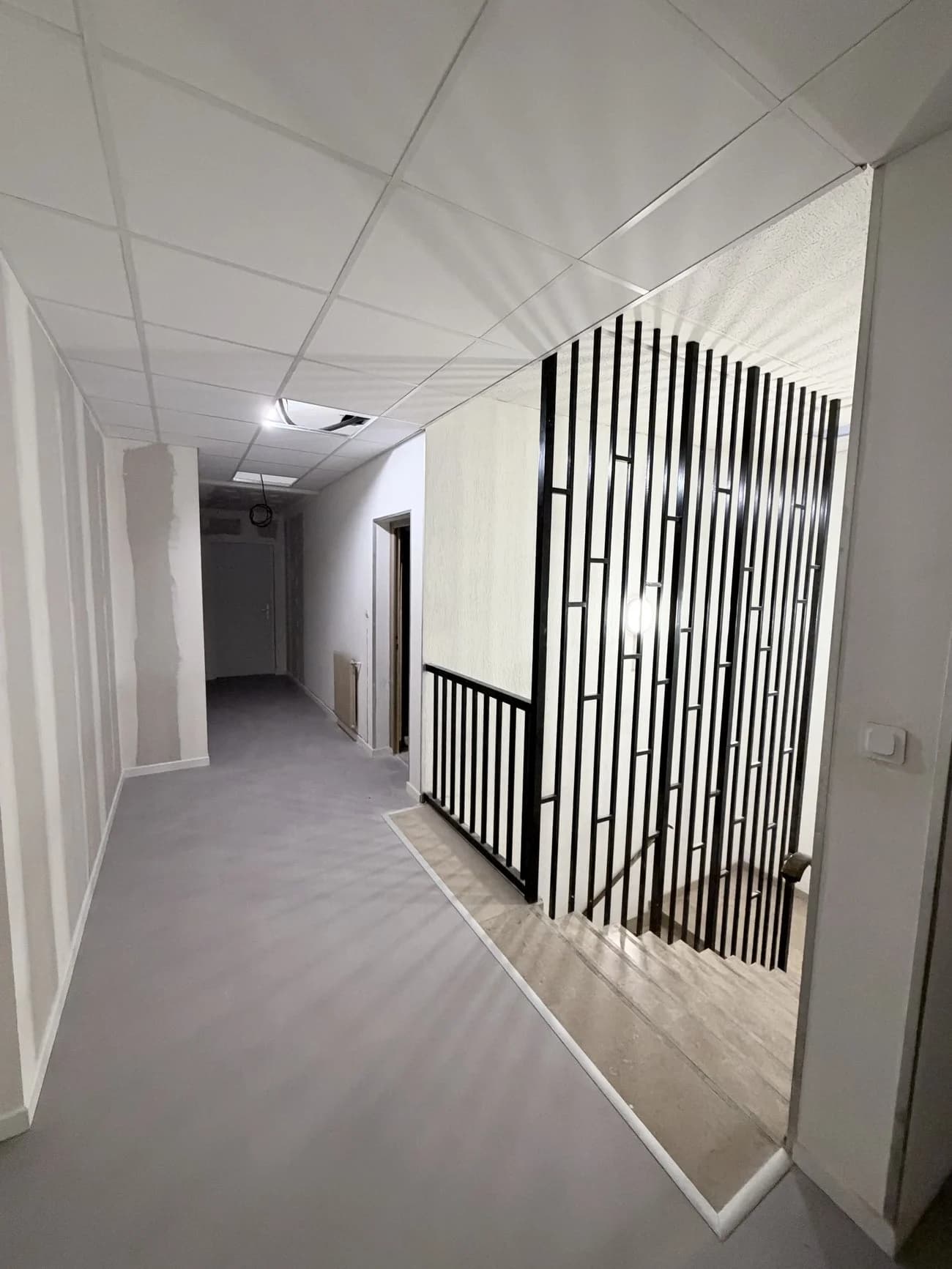 Circulation commune d’un immeuble rénovée avec sol gris, murs clairs, faux plafond et séparation à lames verticales