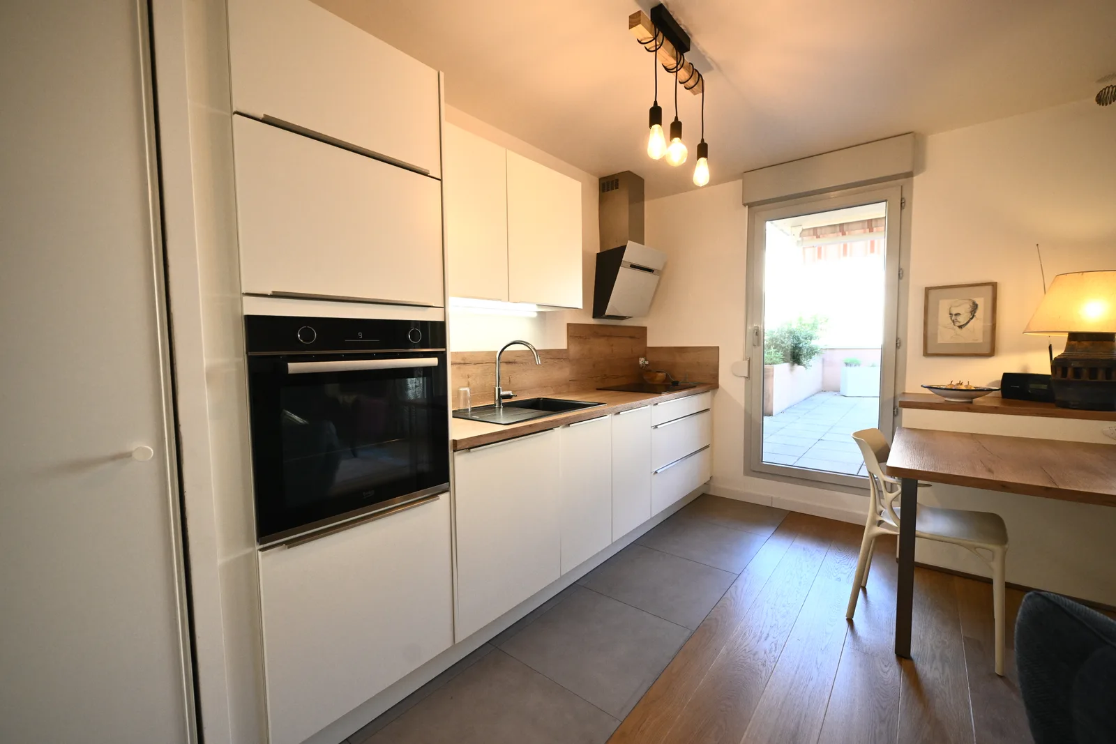Cuisine d’appartement rénovée avec façades blanches, plan de travail en bois et accès à la terrasse